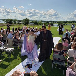 May include: A wedding ceremony taking place outdoors with the bride and groom walking down the aisle. The bride is wearing a white and purple dress and the groom is wearing a black robe. The aisle is decorated with white petals.