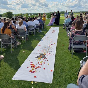 May include: An outdoor wedding ceremony with a white aisle runner decorated with red, yellow, and orange flower petals. The aisle runner has a printed poem on it. The guests are seated in chairs on the green grass.