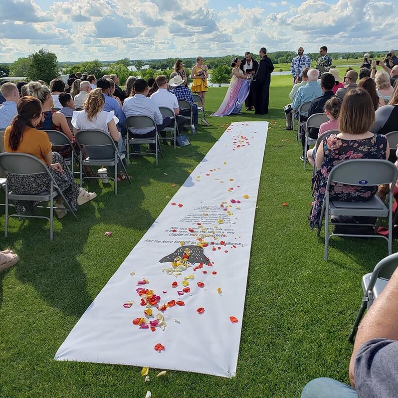 May include: An outdoor wedding ceremony with a white aisle runner decorated with red, yellow, and orange flower petals. The aisle runner has a printed poem on it. The guests are seated in chairs on the green grass.