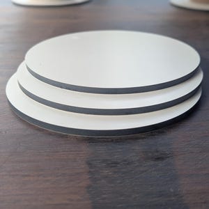 May include: Three round white coasters with black edges are stacked on a dark wooden surface. The coasters are made of a smooth material. The coasters are designed to protect surfaces from drink rings.