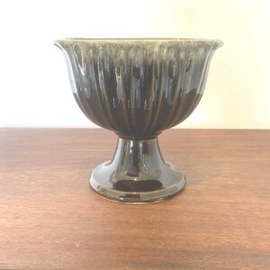 May include: A dark brown ceramic pedestal bowl with a fluted exterior. The bowl has a wide, flared rim and sits on a short, rounded pedestal base. The bowl is approximately 8 inches tall.
