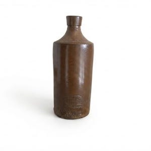 May include: A brown stoneware bottle with a cylindrical body and a narrow neck. The surface has a smooth, glazed finish. Near the base, there is an embossed stamp featuring the text "W. H. & H. LE MAY" and "LONDON" within a curved border.