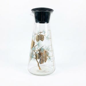 May include: A clear glass pitcher with a black plastic lid, featuring a decorative print of gold pinecones and light blue pine needles. The side of the pitcher displays vertical measurement markings in gold and the word DOUGLAS printed near the base.