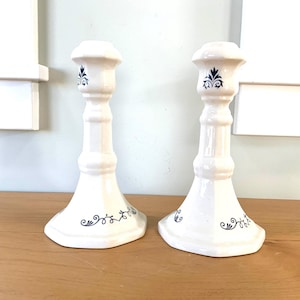 May include: Two off-white ceramic candlesticks with a classic design. Each candlestick features a flared base, a slender stem, and a top with a decorative blue floral motif. The candlesticks are displayed on a wooden surface.