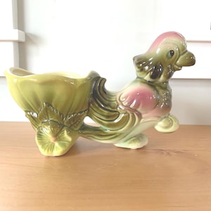 May include: A green and pink ceramic planter shaped like a bird pulling a wagon. The wagon is a flower pot with a green and white floral design.
