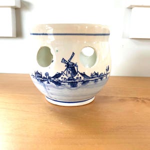 May include: A ceramic, bulbous container with three circular openings. The off-white surface is decorated with a blue and white windmill scene. The rim and base feature blue stripes. The container sits on a wooden surface.