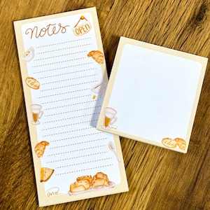 May include: Two beige notepads with a bakery theme. One notepad has the word "Notes" and "OPEN" written on it, with illustrations of pastries and coffee. The other notepad is blank. Both have a light brown border.