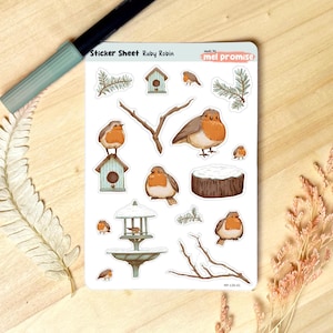 Peut inclure: Une planche d'autocollants intitulée "Sticker Sheet Ruby Robin" avec des illustrations de rouges-gorges, de nichoirs, de branches et d'une mangeoire à oiseaux. Les rouges-gorges ont la poitrine orange et les ailes marron. Les nichoirs sont bleu clair et vert.