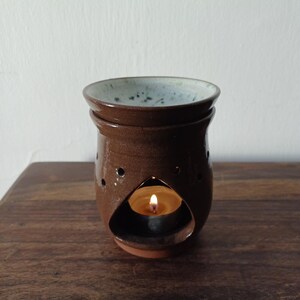 Può includere: Un bruciatore per oli in ceramica marrone con un top smaltato bianco e una piccola candela all'interno. Il bruciatore ha un'apertura a forma di lacrima nella parte anteriore e piccoli fori sui lati.