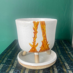 May include: White ceramic planter with a raised base and three legs. The planter has a speckled glaze and features a hand-painted design of two orange-brown trees.