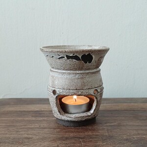 Może przedstawiać: Ceramiczny dyfuzor olejków eterycznych z zapaloną świeczką typu tealight w środku. Dyfuzor jest w jasnoszarym kolorze z teksturowaną powierzchnią i dekoracyjnymi wycięciami. Płomień świecy jest jasny i ciepły.