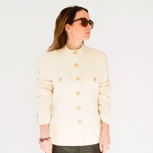K&ouml;nnte beinhalten: Ein wei&szlig;er Blazer mit goldenen Kn&ouml;pfen und einem Stehkragen. Der Blazer hat zwei Pattentaschen auf der Vorderseite.