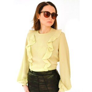 May include: A light yellow blouse with ruffled detailing around the neckline and shoulders. The blouse has long sleeves and a round neckline.