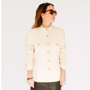 K&ouml;nnte beinhalten: Ein wei&szlig;er Blazer mit goldenen Kn&ouml;pfen und einem Stehkragen. Der Blazer hat eine Patte auf der linken Seite und einen Knopfverschluss.