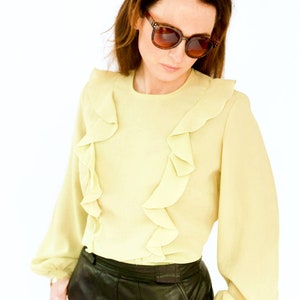 May include: A woman wearing a light yellow blouse with ruffled trim and a black leather skirt. She is wearing brown sunglasses.