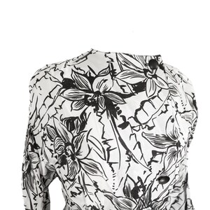 Puede incluir: Una blusa blanca de manga larga con estampado floral negro. La blusa tiene un cuello alto y un cierre de bot&oacute;n en el cuello.