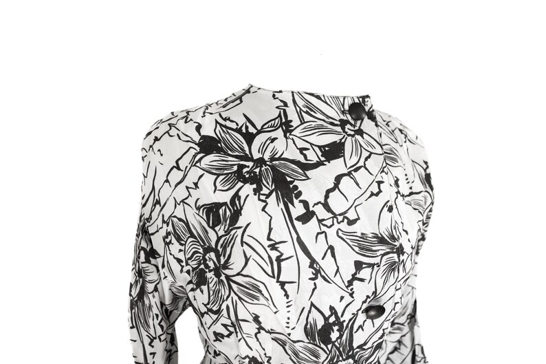 Puede incluir: Una blusa blanca de manga larga con estampado floral negro. La blusa tiene un cuello alto y un cierre de bot&oacute;n en el cuello.