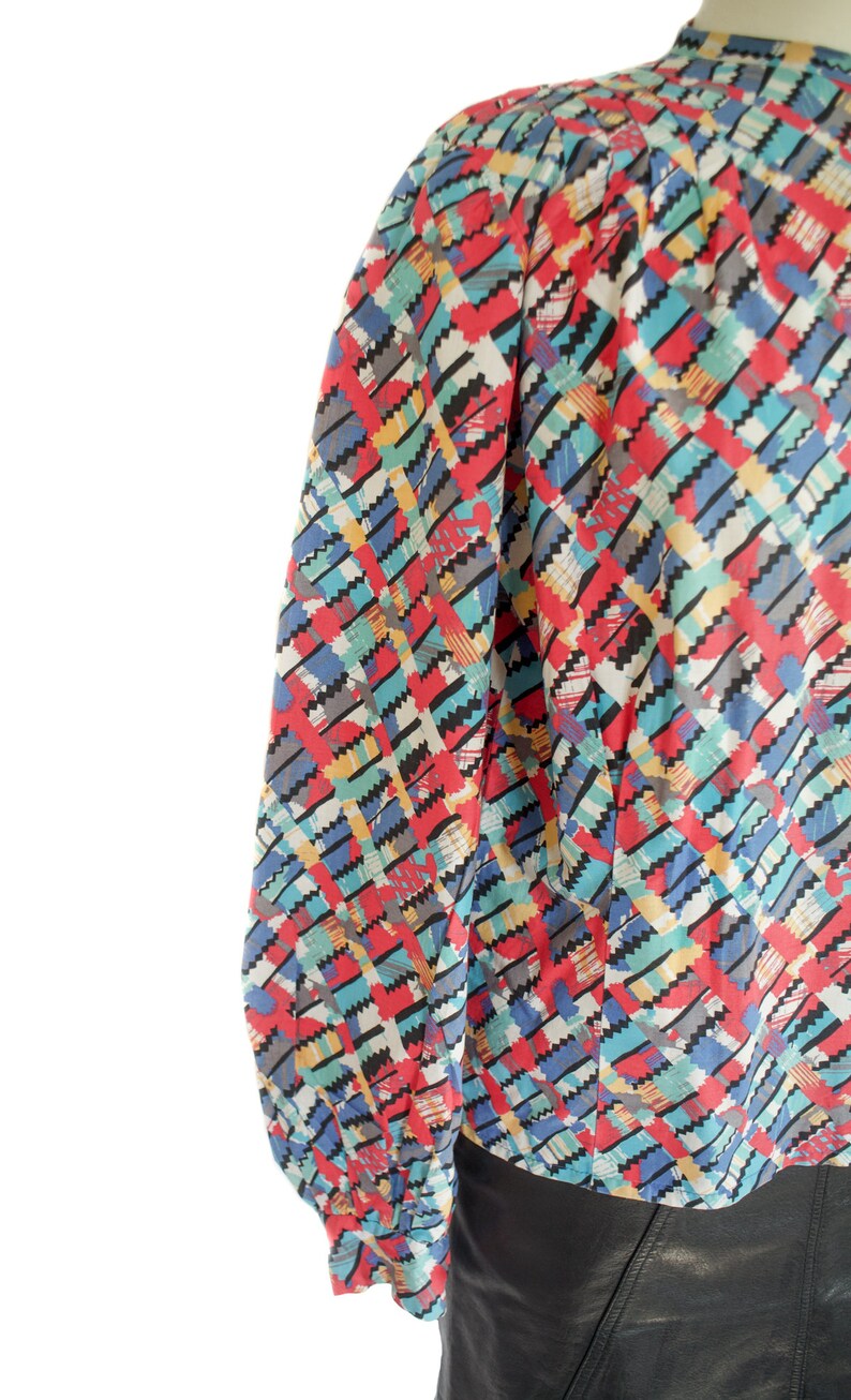 May include: A colorful long-sleeve blouse with a geometric pattern. The pattern features red, blue, yellow, and gray zigzags on a white background. The blouse has a high neckline and gathered sleeves.