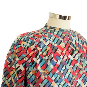 May include: A colorful, patterned blouse with a high neckline. The design features a geometric pattern in red, blue, green, and black. The blouse is displayed on a mannequin.