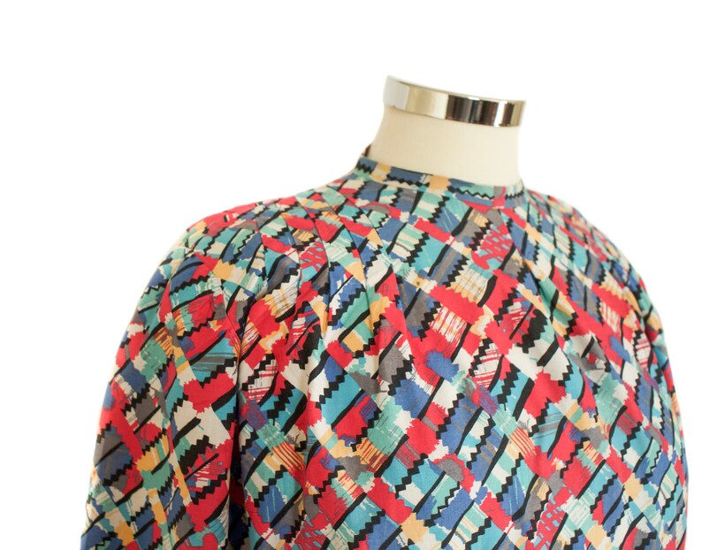 May include: A colorful, patterned blouse with a high neckline. The design features a geometric pattern in red, blue, green, and black. The blouse is displayed on a mannequin.