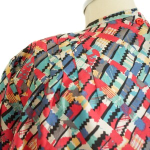 May include: A colorful patterned blouse with a mandarin collar and red buttons. The fabric features a geometric design in red, blue, teal, black, and gray. The blouse is displayed on a mannequin.
