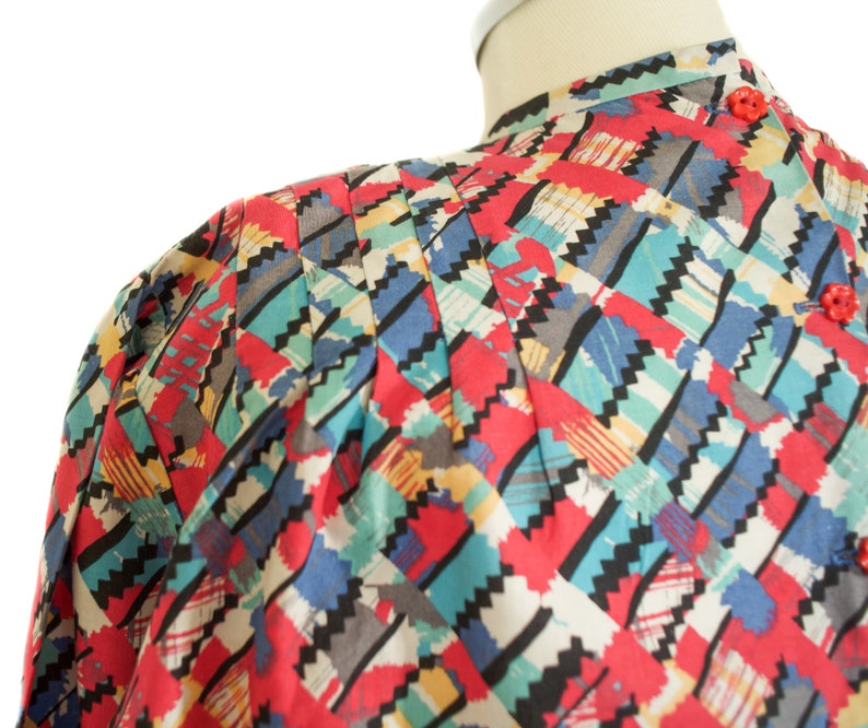 May include: A colorful patterned blouse with a mandarin collar and red buttons. The fabric features a geometric design in red, blue, teal, black, and gray. The blouse is displayed on a mannequin.