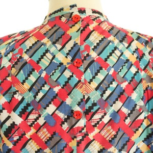 May include: A colorful, patterned blouse with a high collar and red buttons. The fabric features a geometric design with red, blue, teal, yellow, and black zig-zag shapes. The blouse is displayed on a mannequin.