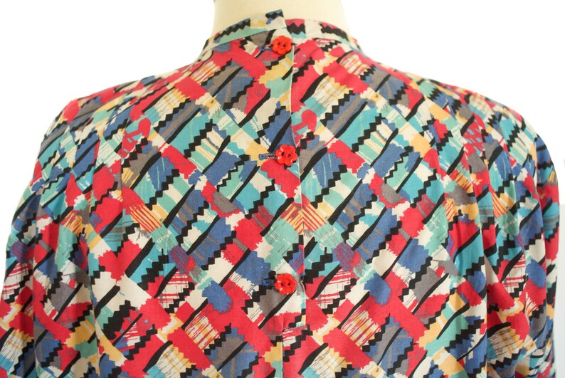 May include: A colorful, patterned blouse with a high collar and red buttons. The fabric features a geometric design with red, blue, teal, yellow, and black zig-zag shapes. The blouse is displayed on a mannequin.