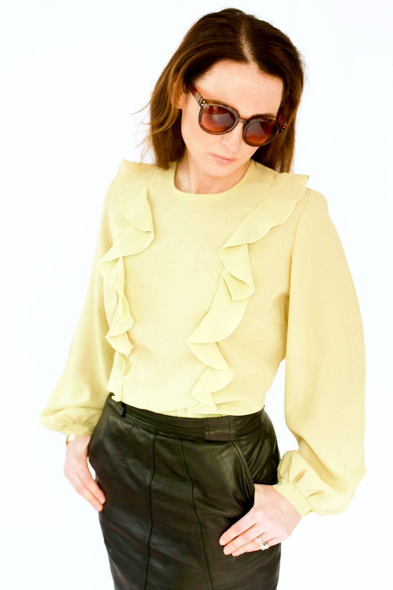 May include: A woman wearing a light yellow blouse with ruffled trim and a black leather skirt. She is wearing brown sunglasses.