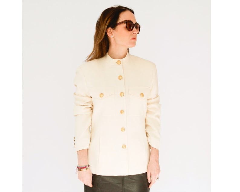 K&ouml;nnte beinhalten: Ein wei&szlig;er Blazer mit goldenen Kn&ouml;pfen und einem Stehkragen. Der Blazer hat eine Patte auf der linken Seite und einen Knopfverschluss.