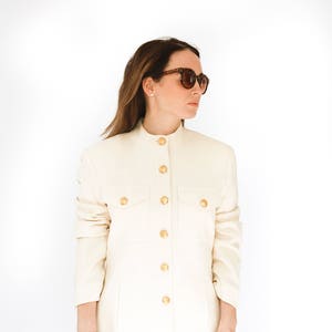 K&ouml;nnte beinhalten: Eine wei&szlig;e Jacke mit langen &Auml;rmeln, goldenen Kn&ouml;pfen und einem Stehkragen. Die Jacke hat zwei gro&szlig;e Taschen auf der Vorderseite. Die Person, die die Jacke tr&auml;gt, tr&auml;gt auch eine braune Sonnenbrille.