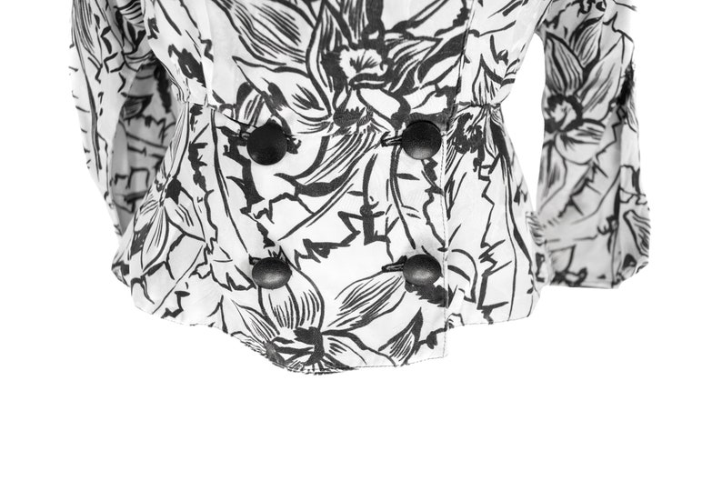 Puede incluir: Una blusa blanca con estampado floral negro y cuatro botones negros en la parte delantera.