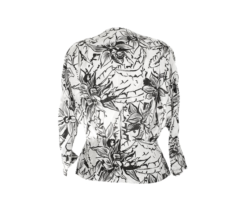 Puede incluir: Una blusa blanca con estampado floral negro. El estampado presenta flores y hojas grandes y estilizadas.
