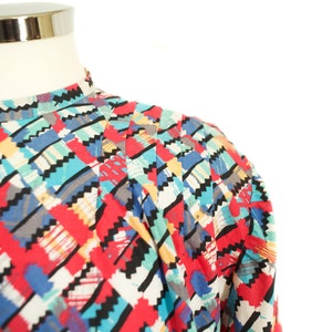 May include: A colorful patterned top with a high collar. The fabric features a geometric design with red, blue, black, and white shapes. The top is displayed on a mannequin, showcasing the shoulder and upper chest area.