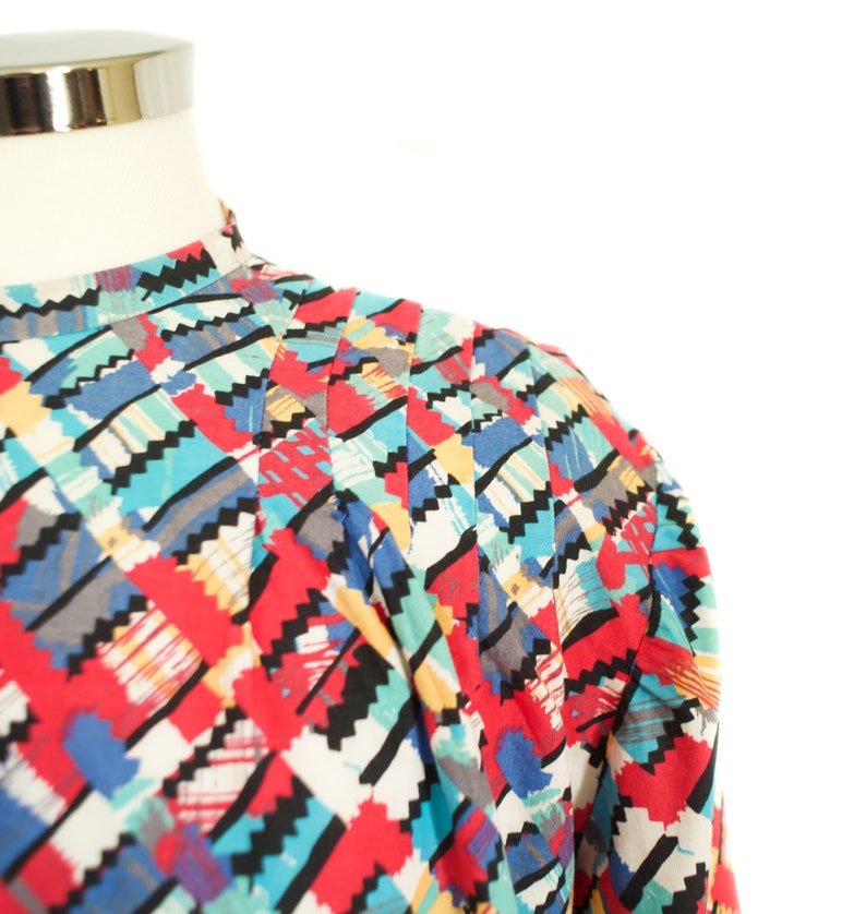 May include: A colorful patterned top with a high collar. The fabric features a geometric design with red, blue, black, and white shapes. The top is displayed on a mannequin, showcasing the shoulder and upper chest area.