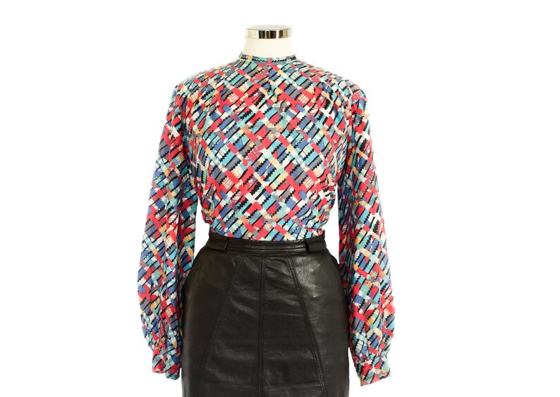 May include: A long-sleeved blouse with a high neckline and a colorful geometric pattern in red, blue, and black. The blouse is paired with a black leather skirt, creating a vintage look.