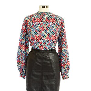 May include: A long-sleeved blouse with a colorful geometric pattern in red, blue, black, and white. The blouse is paired with a black leather skirt. The blouse has a high neckline and the skirt has a fitted waist.