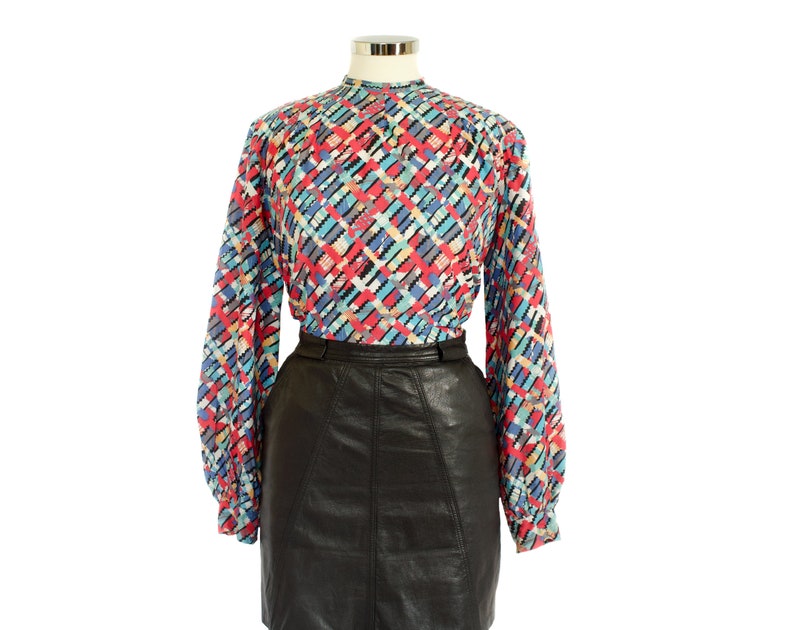 May include: A long-sleeved blouse with a colorful geometric pattern in red, blue, black, and white. The blouse is paired with a black leather skirt. The blouse has a high neckline and the skirt has a fitted waist.