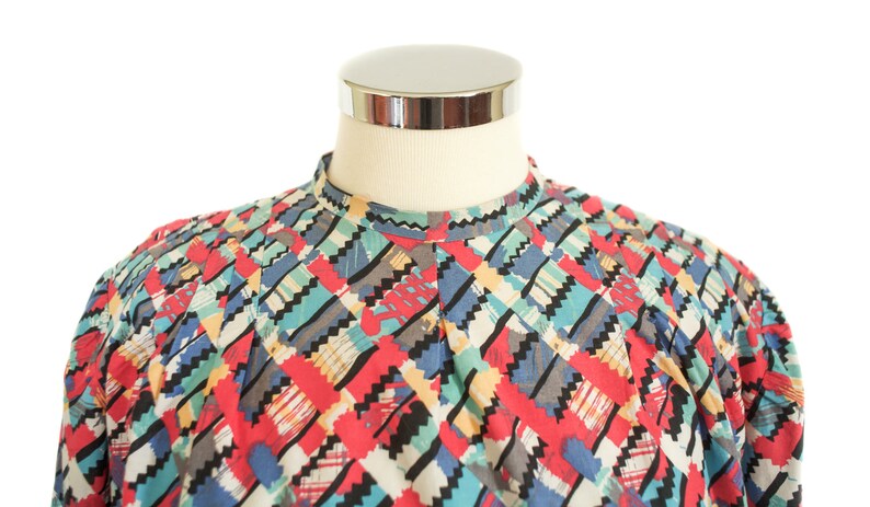 May include: A colorful patterned shirt with a high collar. The shirt features a geometric design with red, blue, black, gray, and yellow accents. The shirt is displayed on a mannequin.