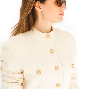 K&ouml;nnte beinhalten: Ein wei&szlig;er Blazer mit goldenen Kn&ouml;pfen und einem Stehkragen. Der Blazer hat eine Tasche auf der linken Seite und ist aus glattem Stoff gefertigt.