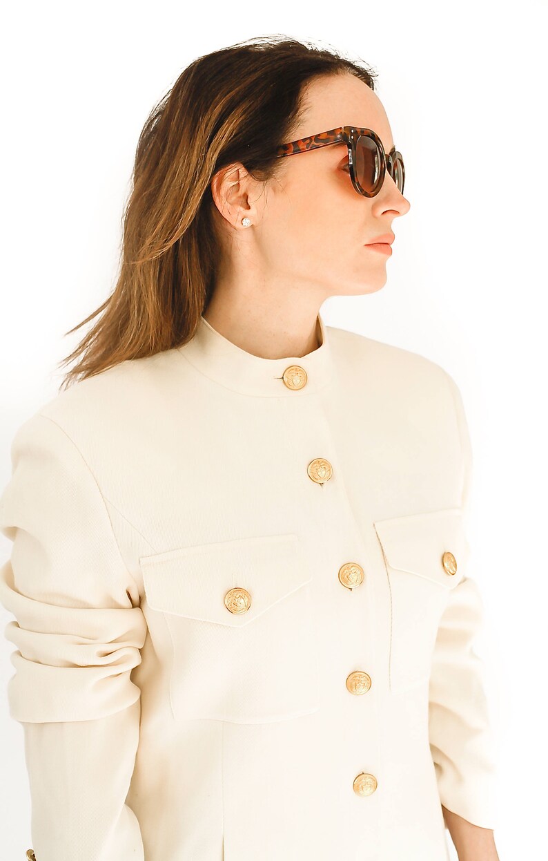 K&ouml;nnte beinhalten: Ein wei&szlig;er Blazer mit goldenen Kn&ouml;pfen und einem Stehkragen. Der Blazer hat eine Tasche auf der linken Seite und ist aus glattem Stoff gefertigt.