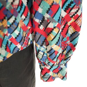 May include: A colorful patterned blouse with long sleeves. The fabric features a geometric design in red, blue, yellow, and gray. The sleeves are gathered at the wrist with an elastic cuff. The blouse is paired with a black garment.