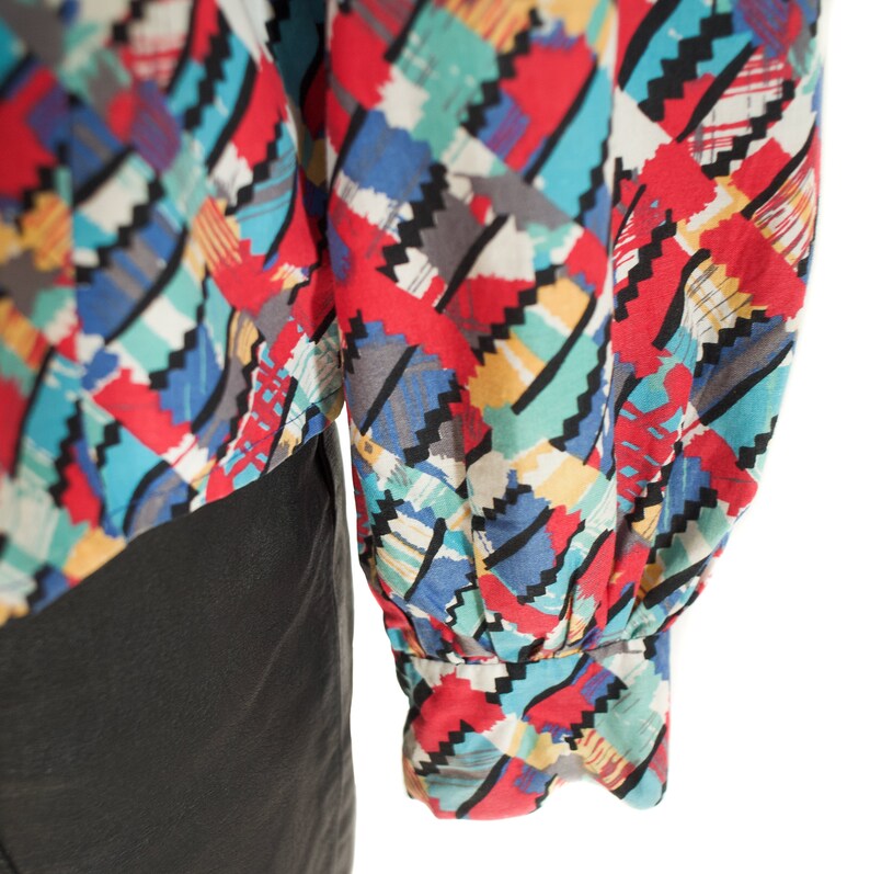 May include: A colorful patterned blouse with long sleeves. The fabric features a geometric design in red, blue, yellow, and gray. The sleeves are gathered at the wrist with an elastic cuff. The blouse is paired with a black garment.