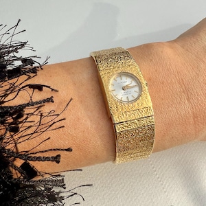 May include: A gold wristwatch with a textured strap and a round face. The watch face is white with black hands and the text "HUNTANA" and "17 JEWELS INCABLOC". The strap is wide and wraps around the wrist.