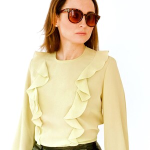 May include: A woman wearing a light green blouse with ruffles and a black leather skirt. She is wearing brown tortoise shell sunglasses.