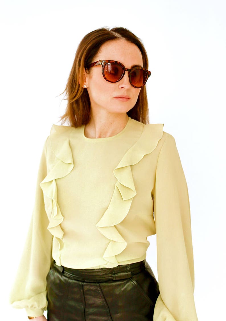 May include: A woman wearing a light green blouse with ruffles and a black leather skirt. She is wearing brown tortoise shell sunglasses.
