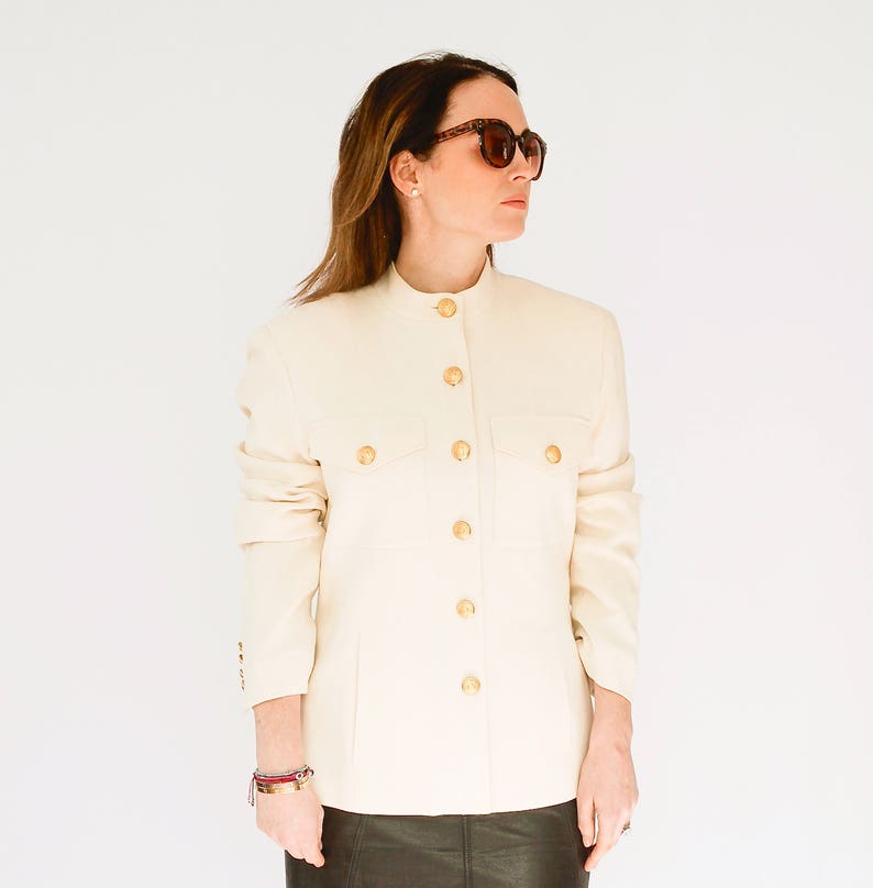 K&ouml;nnte beinhalten: Ein wei&szlig;er Blazer mit goldenen Kn&ouml;pfen und einem Stehkragen. Der Blazer hat zwei Pattentaschen auf der Vorderseite.