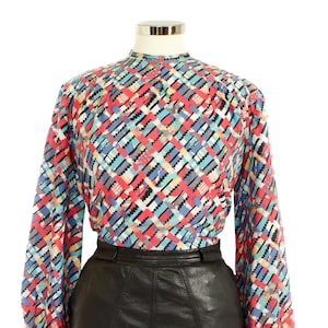 May include: A long-sleeved blouse with a colorful geometric pattern in red, blue, black, and white. The blouse has a high neckline and is paired with a black leather skirt. The top is displayed on a mannequin.