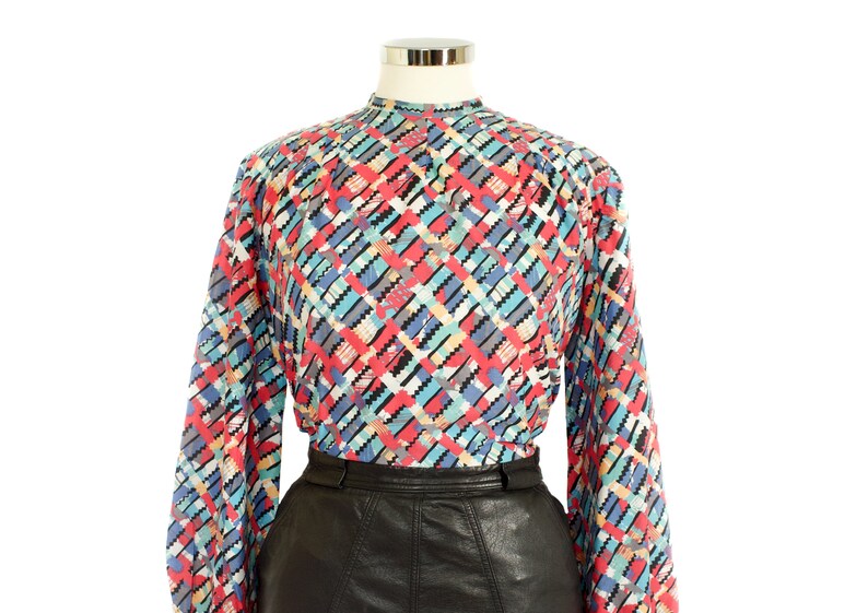 May include: A long-sleeved blouse with a colorful geometric pattern in red, blue, black, and white. The blouse has a high neckline and is paired with a black leather skirt. The top is displayed on a mannequin.