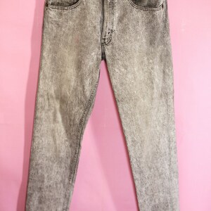 May include: Grey acid wash jeans with a straight leg fit. The jeans are hanging on a black hanger against a pink background.